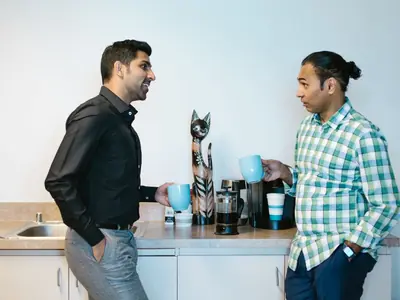 Dos personas conversando en la cocina de una oficina junto a la cafetera